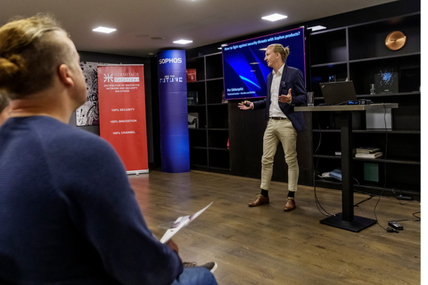 Presenter speaking in front of an audience wearing a Catchbox Clip microphone for audio. A screen behind displays a presentation, and branded banners are visible in the background.