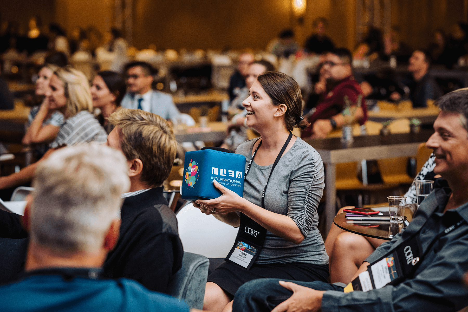 Smiling attendee holding a customized Catchbox Cube microphone during an interactive session at a conference, with other engaged participants seated in the background.