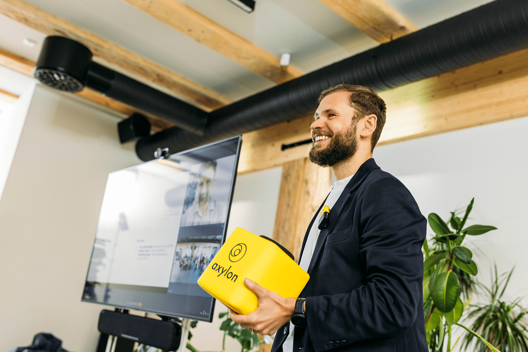 A smiling presenter holding a yellow Catchbox Cube microphone during a hybrid meeting, with a screen in the background displaying remote participants