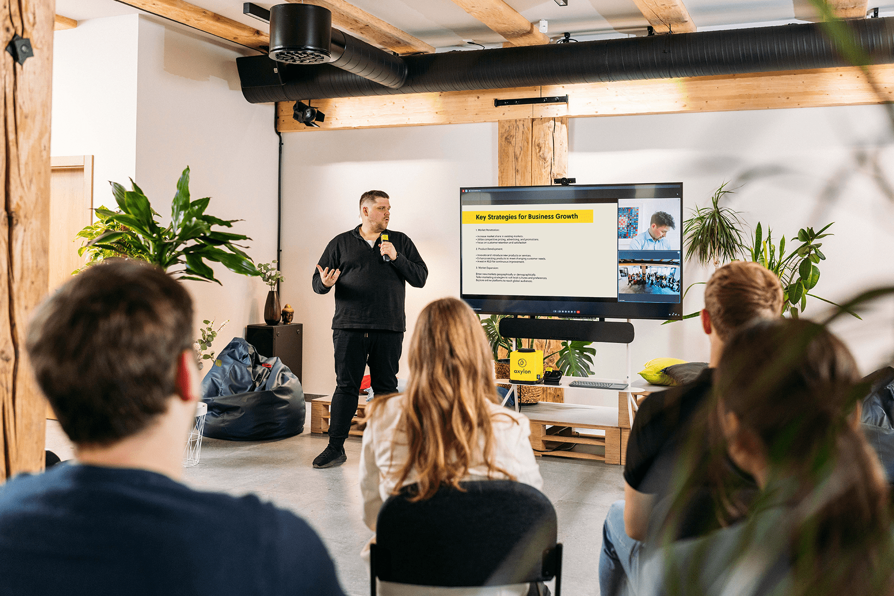 Presenter delivering a session and speaking into a Catchbox Stick handheld microphone, with a screen displaying presentation slides and virtual attendees in the background.