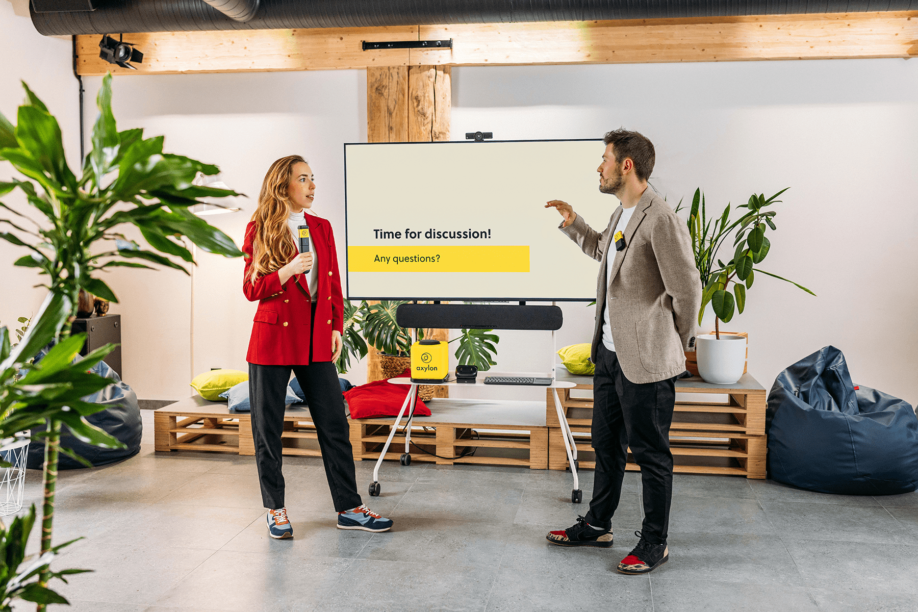 Two presenters standing in front of a screen displaying 'Time for discussion! Any questions?' The woman on the left holds a Stick handheld microphone, while the man on the right has clipped Clip microphone and gestures toward the screen.