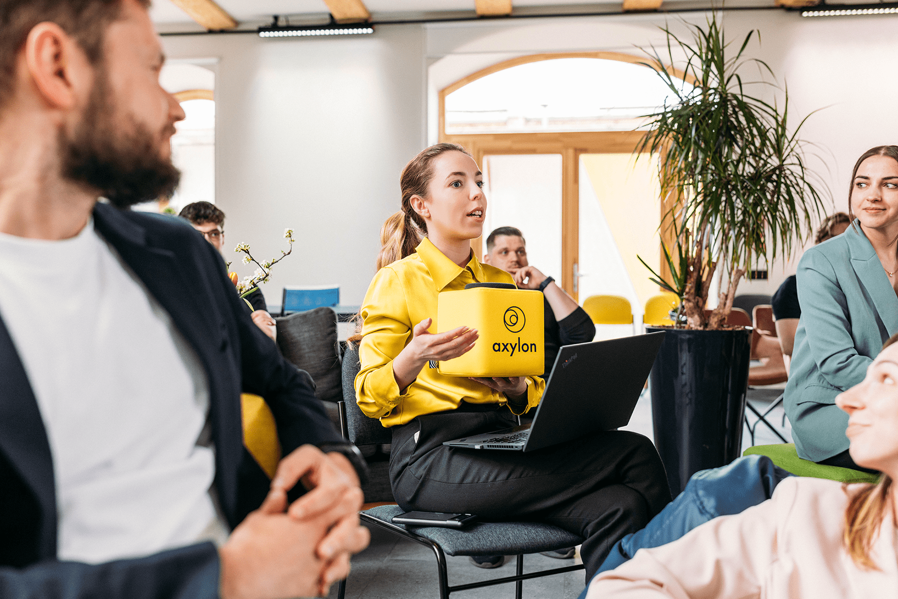 A woman holding a yellow Catchbox Cube microphone while speaking during a meeting, ensuring her voice is clearly captured for both live attendees and the meeting recording.