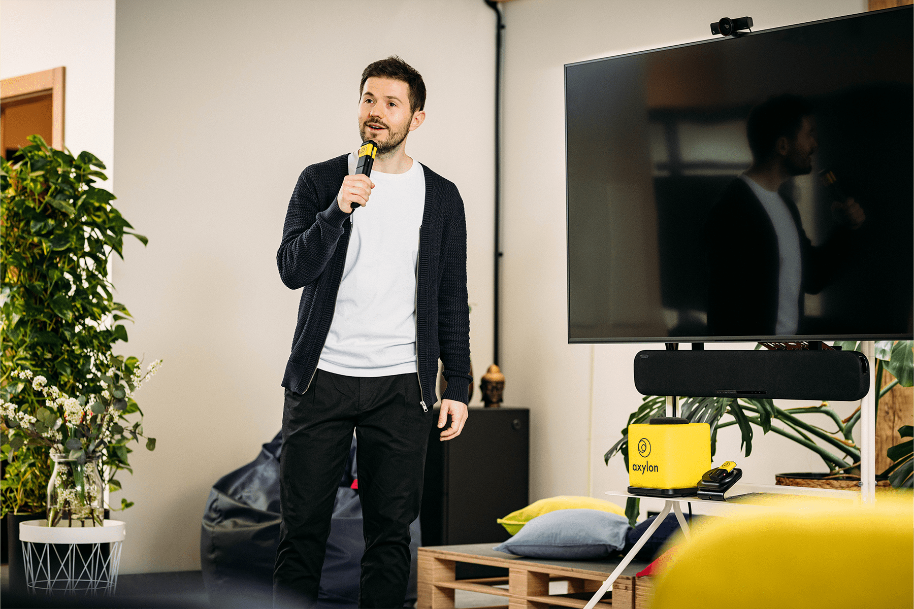 Presenter speaking into a Catchbox Stick handheld microphone during a session, with a screen and a Catchbox Cube and Clip microphones placed in the background.