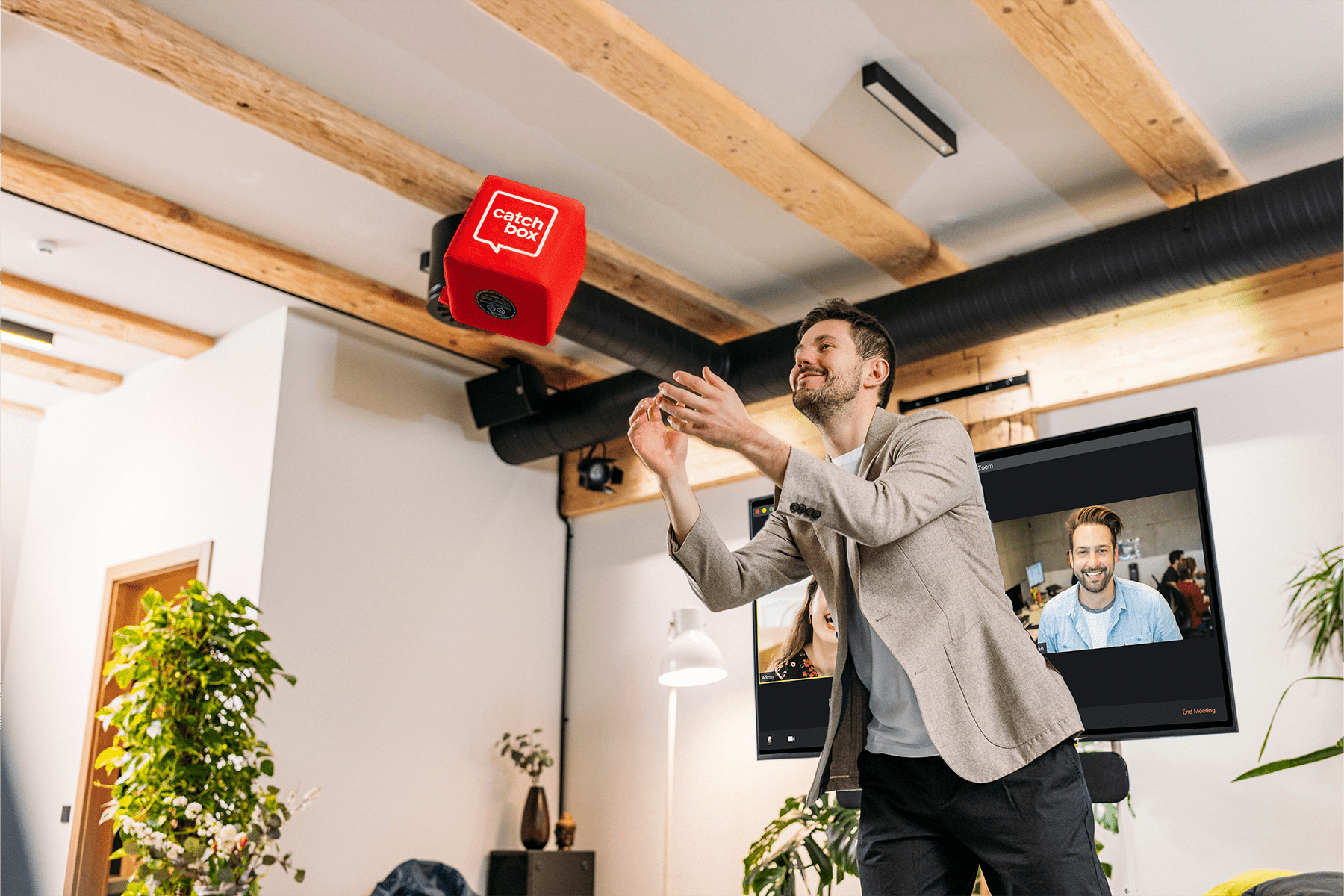 A meeting leader catching a red Catchbox Cube microphone during a hybrid session, with participants visible both in-person and on a video call in the background.